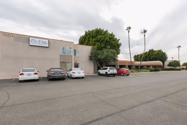 Exterior view of Rio Vista Post Acute And Rehabilitation building with a parking lot in front. Several cars are parked near the entrance, and there are tall palm trees and other greenery around the building under a cloudy sky.