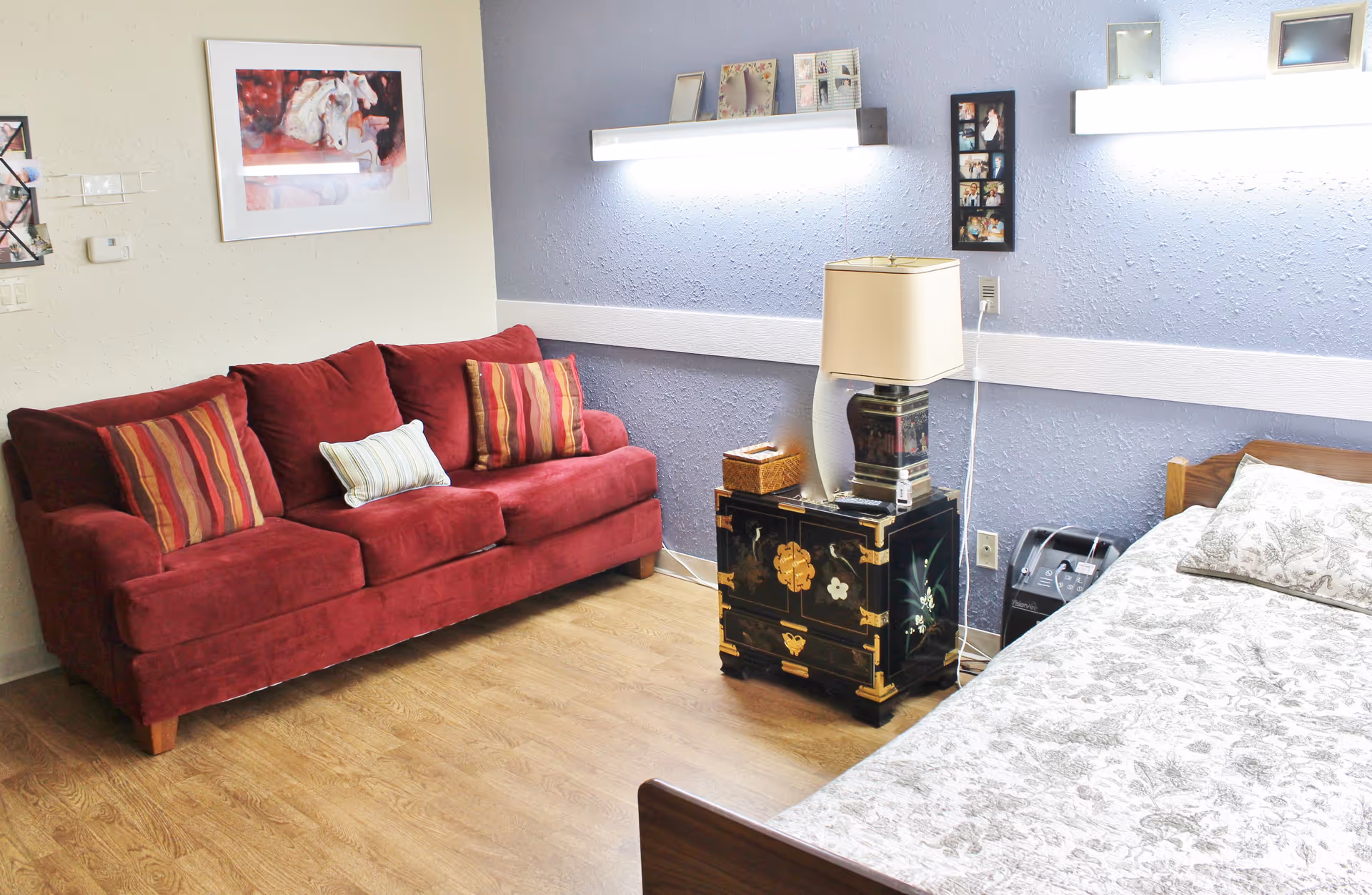 A furnished senior living bedroom with a red sofa, a bed with patterned bedding, and a bedside table with a lamp against a light blue wall.