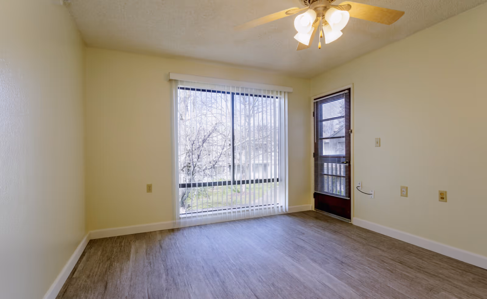 Empty living room with wood-look flooring, a large sliding glass window with vertical blinds, a side door with blinds, and a ceiling fan.