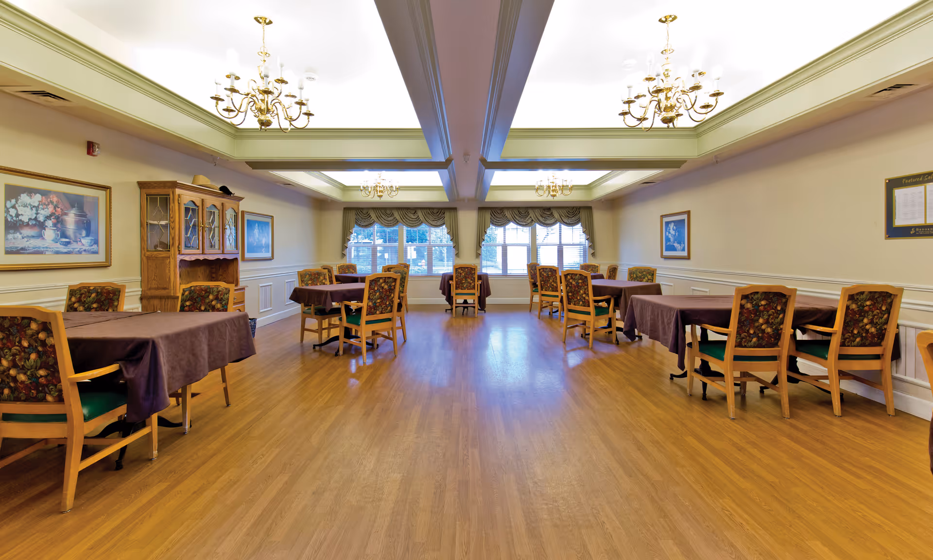 Spacious dining room with multiple tables and floral-upholstered chairs under chandeliers and large windows.