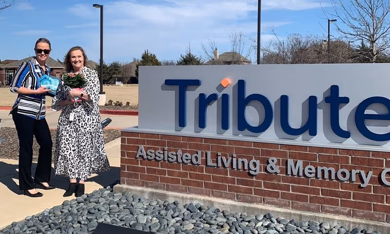 Two women standing outside next to a large sign that reads 'Tribute Assisted Living & Memory Care'. One woman is holding a bouquet of flowers and the other is holding a small blue item. They are smiling and the background shows a clear sky and some trees.