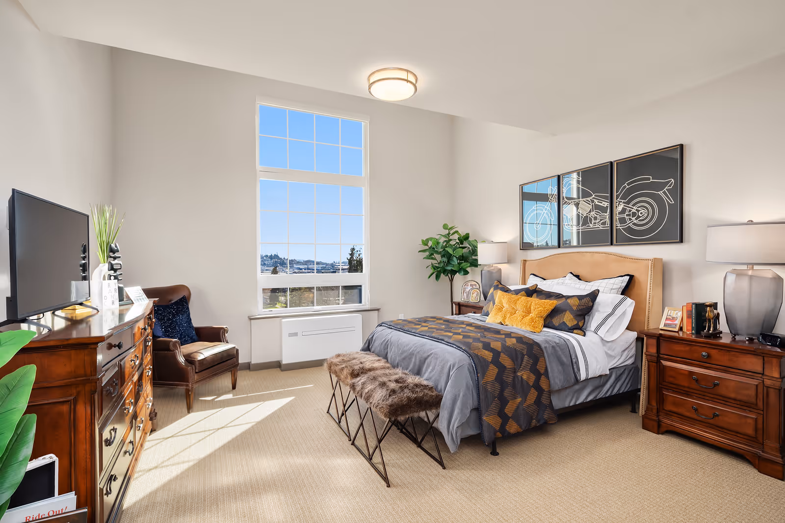 A bright and spacious bedroom with a large window letting in natural light. The room features a neatly made bed with gray and patterned bedding, yellow accent pillows, and a tan upholstered headboard. Above the bed are three framed artworks depicting a motorcycle. On either side of the bed are wooden nightstands with lamps and decorative items. A wooden dresser with a TV on top and a leather armchair with a dark blue pillow are positioned near the window. The room has beige carpeting and a small bench with fur cushions at the foot of the bed.