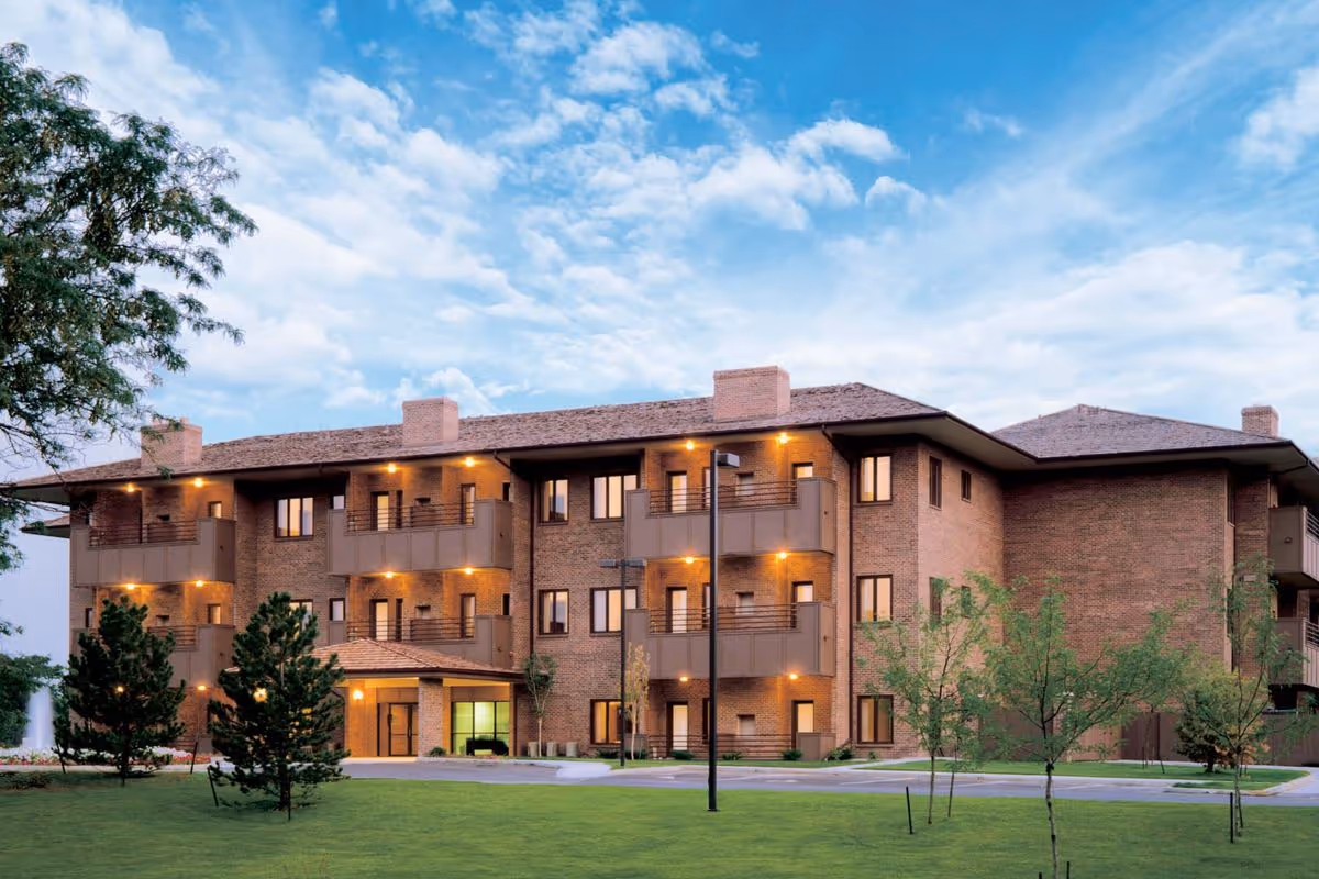 Exterior view of a three-story brick senior living facility building with balconies and warm lighting at dusk, surrounded by a well-maintained lawn and trees under a partly cloudy sky.