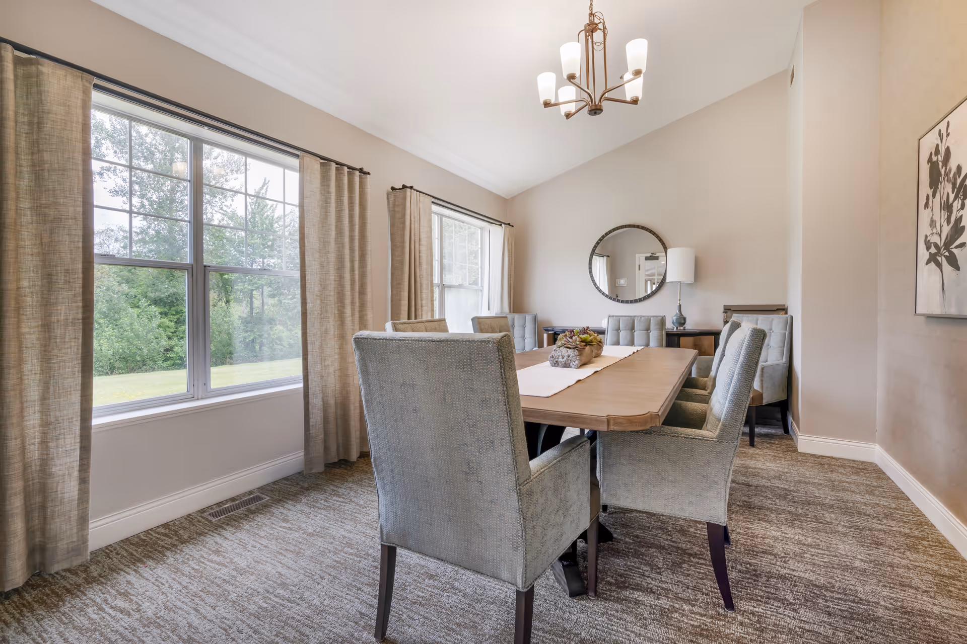 A dining room with a wooden table surrounded by six upholstered chairs. The room has large windows with beige curtains letting in natural light, a round mirror on the wall, a table lamp, and a piece of wall art with a floral design. The carpet is a neutral tone and the walls are painted light beige.
