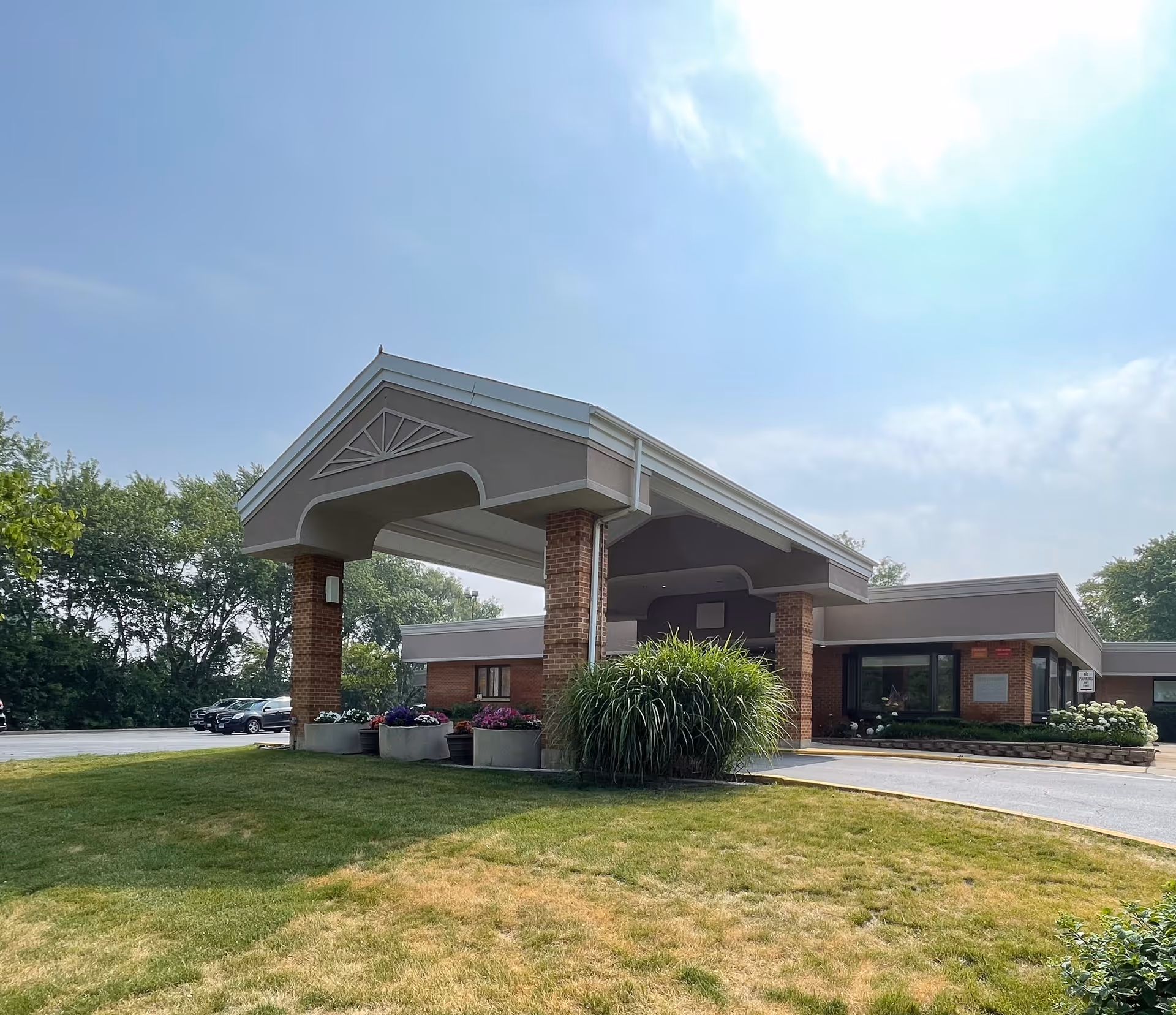 Front entrance of a single-story brick senior living facility with a covered porte-cochère, landscaping, and parked cars.