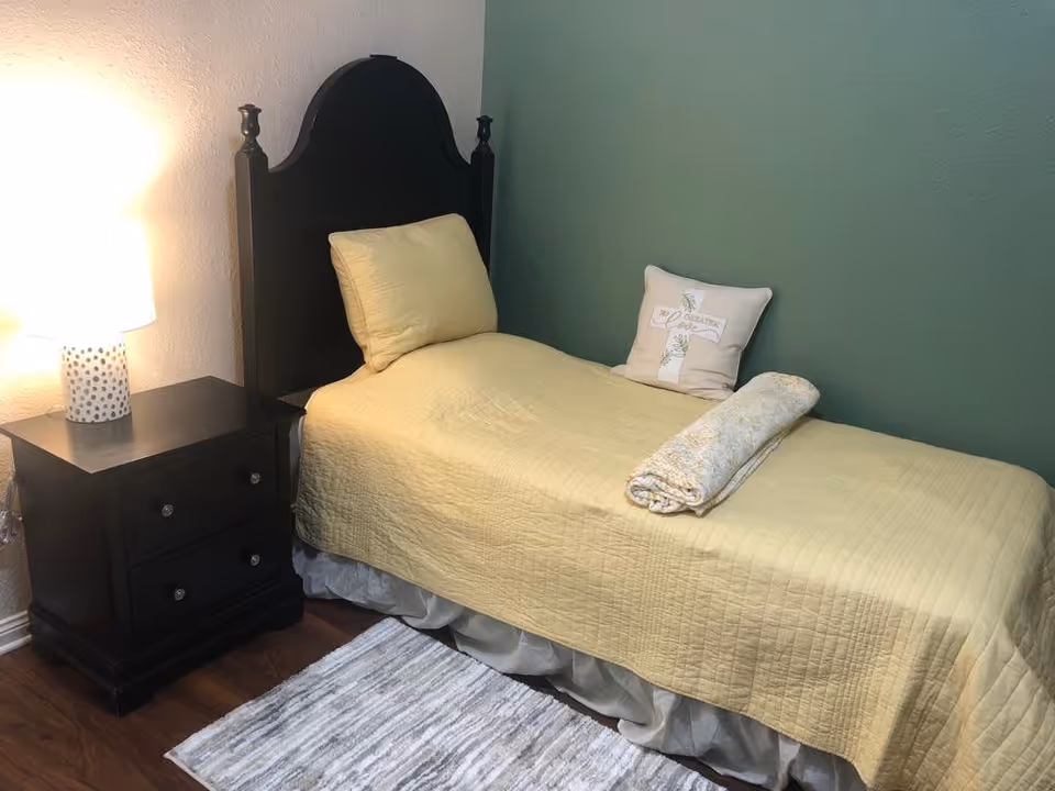 A small bedroom with a single bed covered in a yellow quilt and matching pillow. There is a rolled-up blanket and a decorative pillow on the bed. Next to the bed is a dark wooden nightstand with two drawers and a white lamp with a dotted pattern. The walls are painted green and beige, and there is a small gray and white rug on the wooden floor.