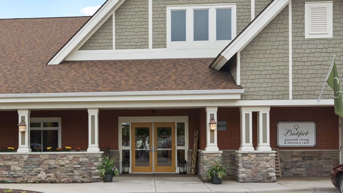 Front entrance of a senior living facility with double glass doors, stone and brick exterior, a green and beige upper facade, two lantern-style lights on columns, potted plants, and a sign that reads 'Bickford assisted living & memory care'.