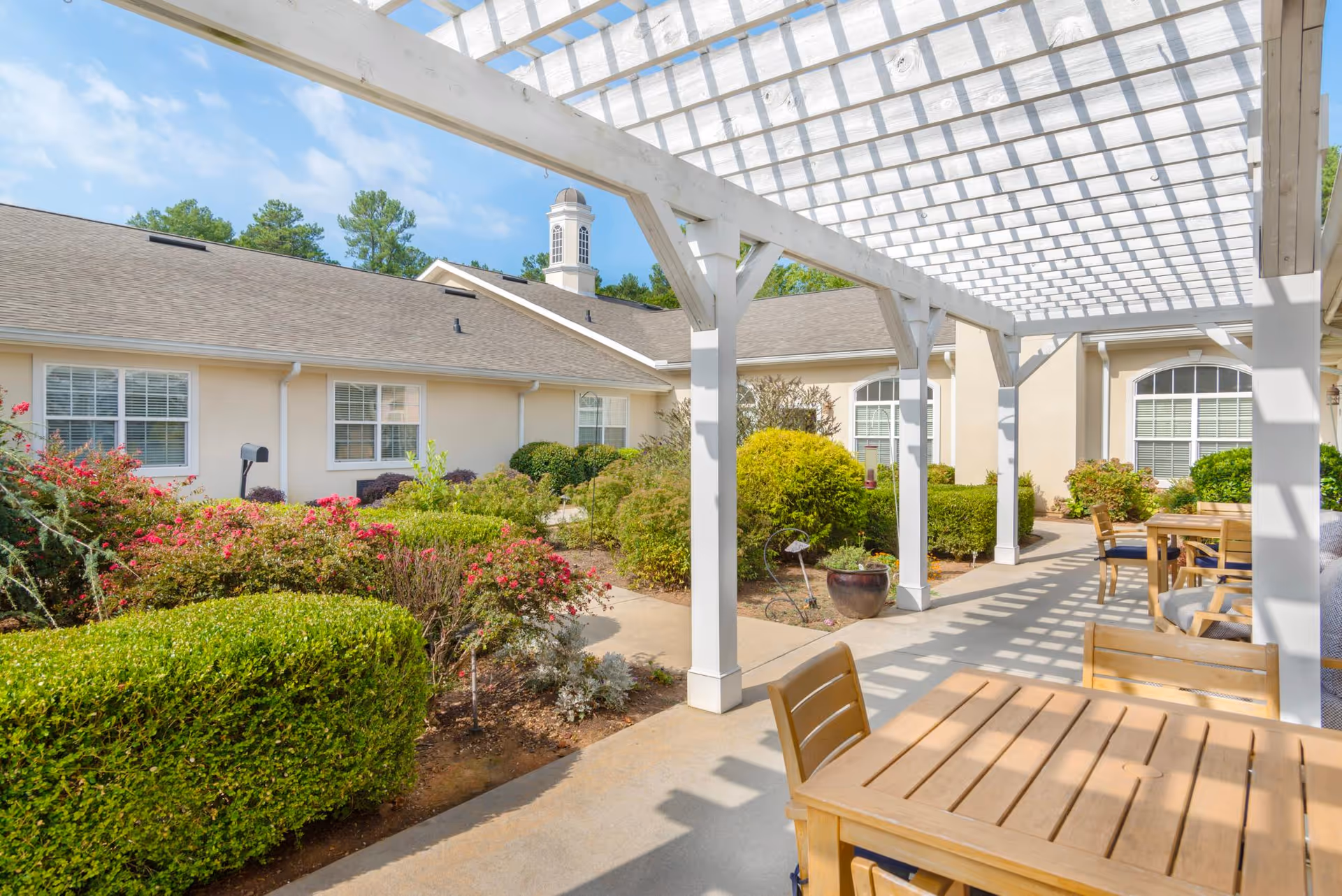 Sunlit courtyard with a white pergola, wooden tables and chairs, and landscaped bushes in front of a single-story senior living building.
