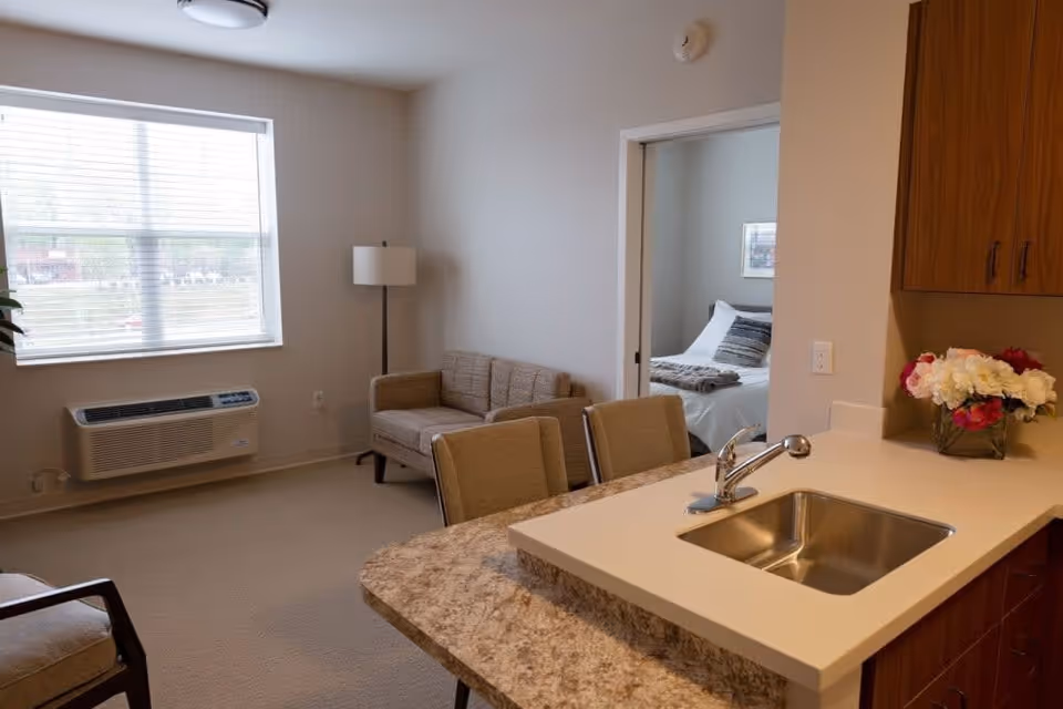 Interior view of a senior living facility apartment showing a small living area with a loveseat, a floor lamp, and a window with blinds. In the foreground, there is a kitchen counter with a sink and a vase of flowers. Through an open doorway, a bedroom with a bed and pillows is visible.