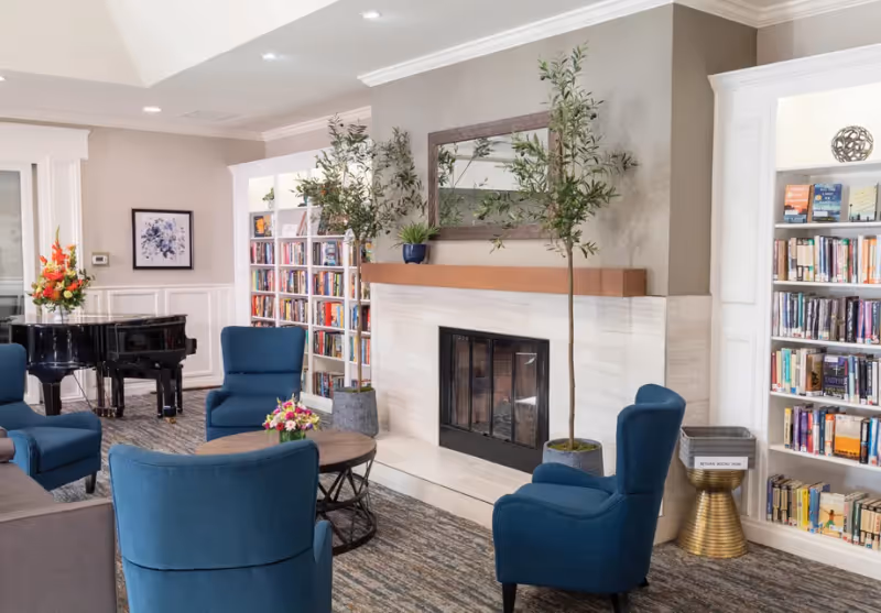A cozy living room area in a senior living facility featuring a fireplace with a wooden mantel, two tall potted plants on either side, a large mirror above the fireplace, blue upholstered armchairs arranged around a round wooden coffee table with a small flower arrangement, bookshelves filled with books, a black grand piano with a flower arrangement on top, and neutral-colored walls and carpet.