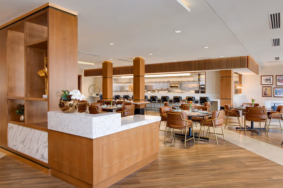 A modern dining room with wooden floors and furniture, featuring several round tables with brown leather chairs. The room has a light and airy atmosphere with a marble countertop in the foreground and an open kitchen area with bar stools in the background. Decorative elements include a vase with white flowers and framed artwork on the walls.