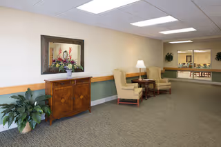 A well-lit interior hallway area with beige walls and carpeted floor. There is a wooden cabinet with a decorative mirror and a flower arrangement on top, a potted plant on the floor to the left, and two beige upholstered armchairs with a small wooden table and lamp between them. In the background, there is a large window opening into another room with tables and chairs.