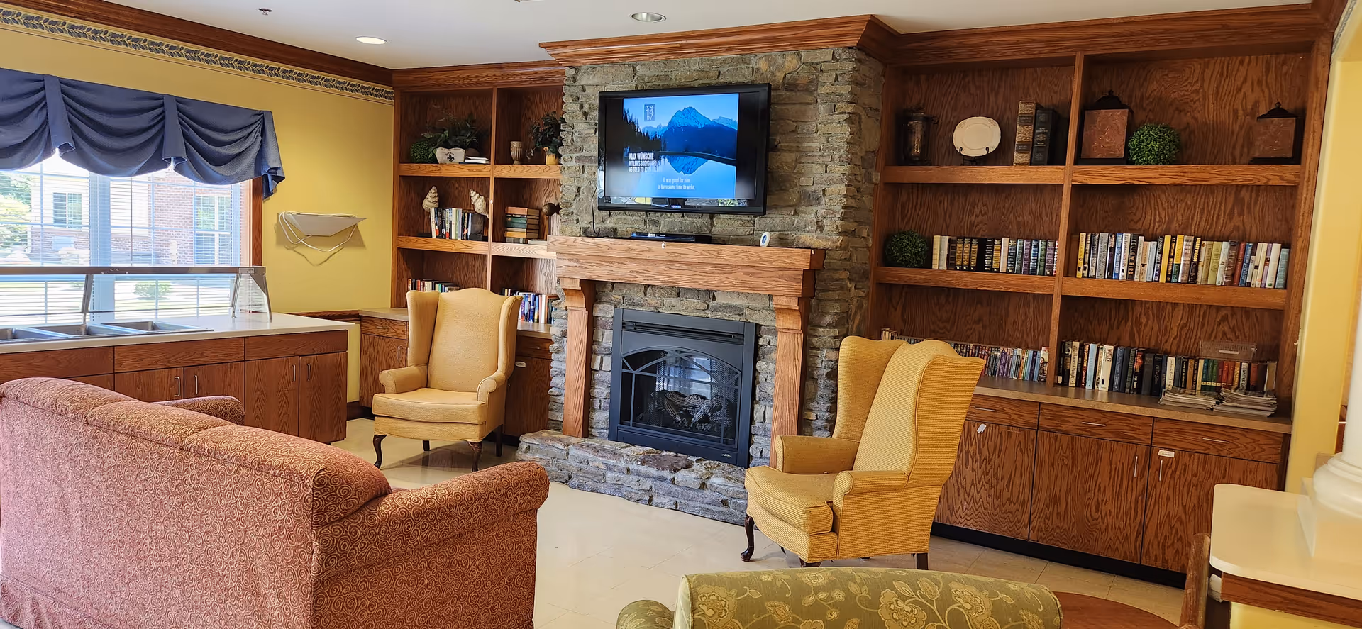 A cozy living room area in a senior living facility featuring a stone fireplace with a mounted flat-screen TV above it. The room includes two yellow upholstered armchairs, a patterned sofa, wooden built-in bookshelves filled with books and decorative items, and a large window with blue valance curtains letting in natural light.
