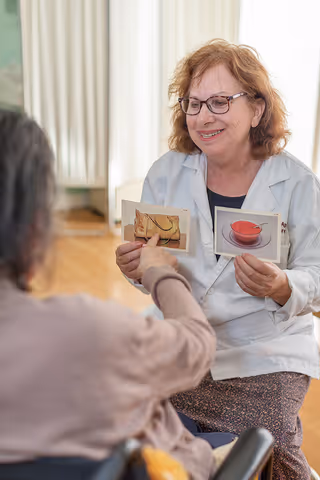 A healthcare professional wearing glasses and a white coat is sitting and smiling while holding two picture cards, one showing a handbag and the other a cup of tea. An elderly person in a wheelchair is pointing at one of the cards in a bright room with wooden flooring and light curtains.