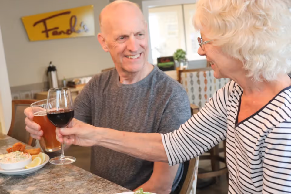 An elderly man and woman sitting at a dining table in a restaurant or dining area, smiling and clinking their glasses in a toast. The man holds a glass of beer and the woman holds a glass of red wine. There is a plate of food on the table and a sign on the wall behind them that reads 'Fanchos'.