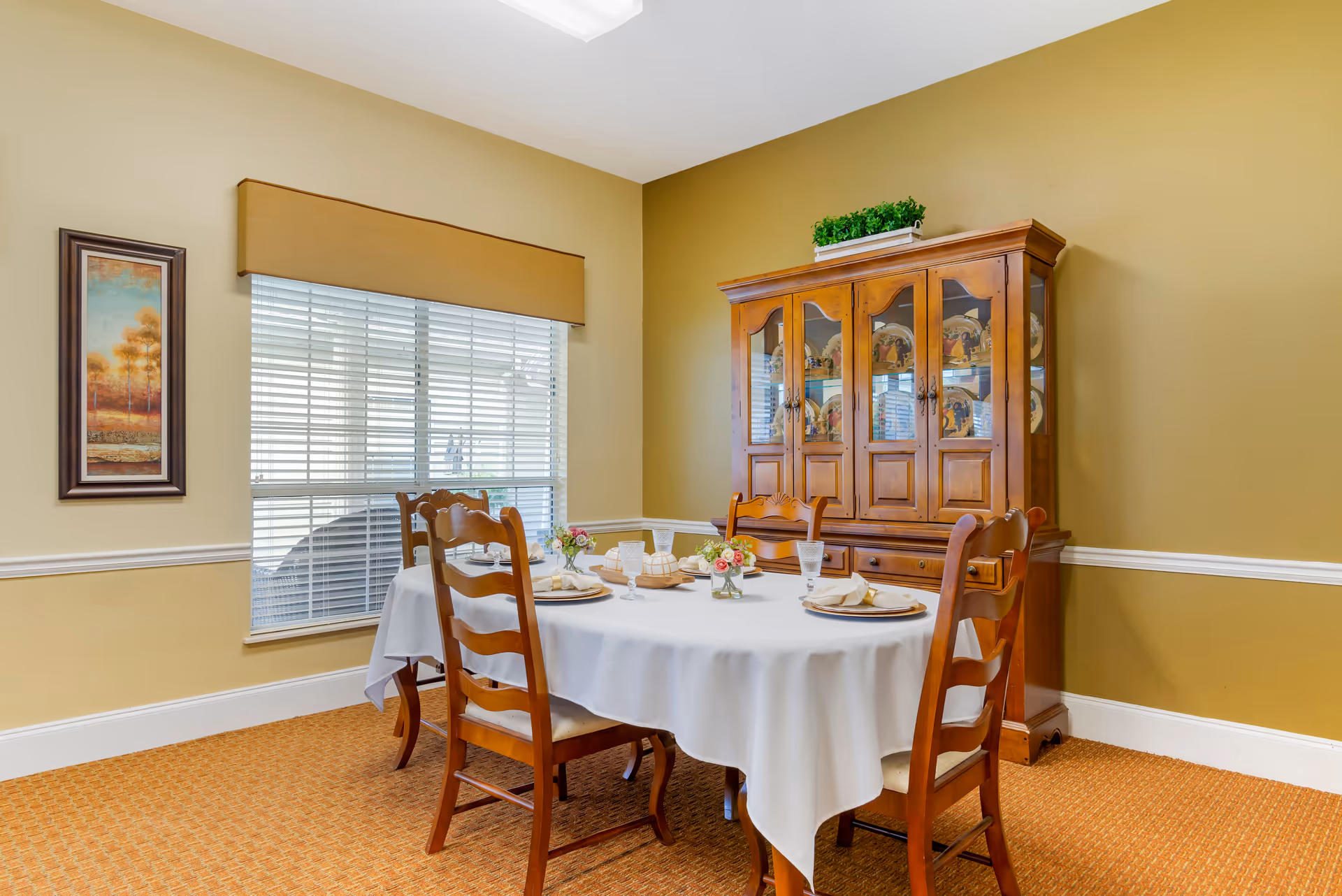 A dining room with a round table covered with a white tablecloth set for four people. The table has plates, napkins, glasses, and small flower arrangements. Behind the table is a wooden china cabinet displaying decorative plates. There is a window with blinds and a valance on the left wall, and a framed painting beside it. The walls are painted in two shades of beige with white trim, and the floor is carpeted in a warm brown pattern.