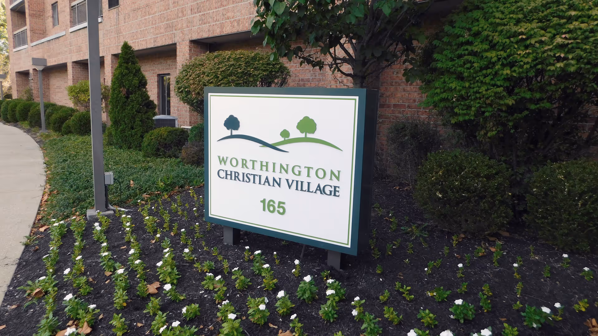 Outdoor view of the Worthington Christian Village sign surrounded by small plants and bushes, with a brick building and sidewalk in the background.