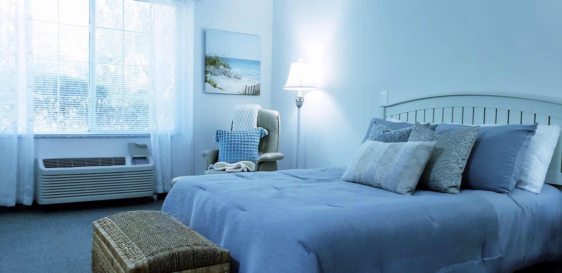 A cozy bedroom with a neatly made bed featuring multiple pillows and a blue comforter. There is a wicker storage bench at the foot of the bed, a comfortable armchair with a blue cushion and a blanket, a floor lamp, and a window with sheer white curtains letting in natural light. A beach-themed painting hangs on the wall.