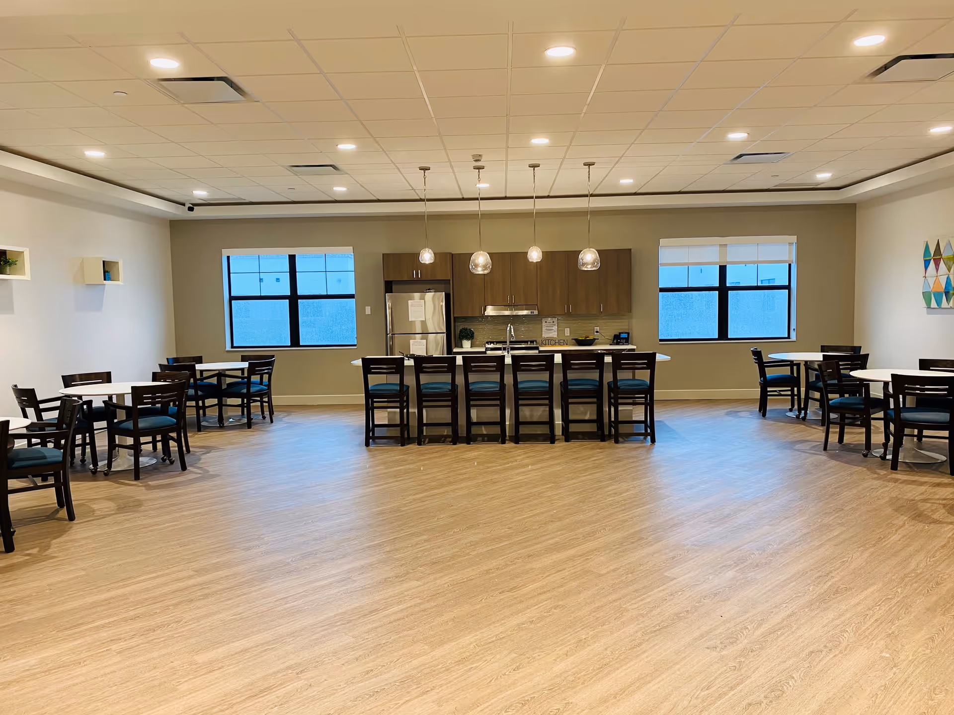 A spacious dining area with light wood flooring and a modern kitchen at the back featuring a stainless steel refrigerator, stove, and wooden cabinets. There are several round tables with dark wooden chairs arranged around the room, and a long island with bar stools in front of the kitchen. The room has two large windows and pendant lights hanging above the island.