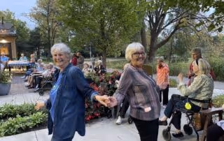 Two elderly women holding hands and smiling outdoors at a senior living facility event, with other seniors sitting and socializing in the background surrounded by trees and greenery.