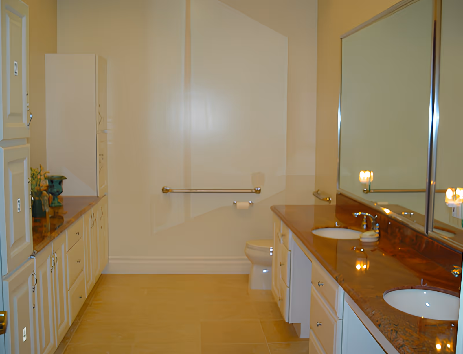 Spacious bathroom with dual sinks set in a long countertop, large mirror, cabinetry, and a toilet with a grab bar.