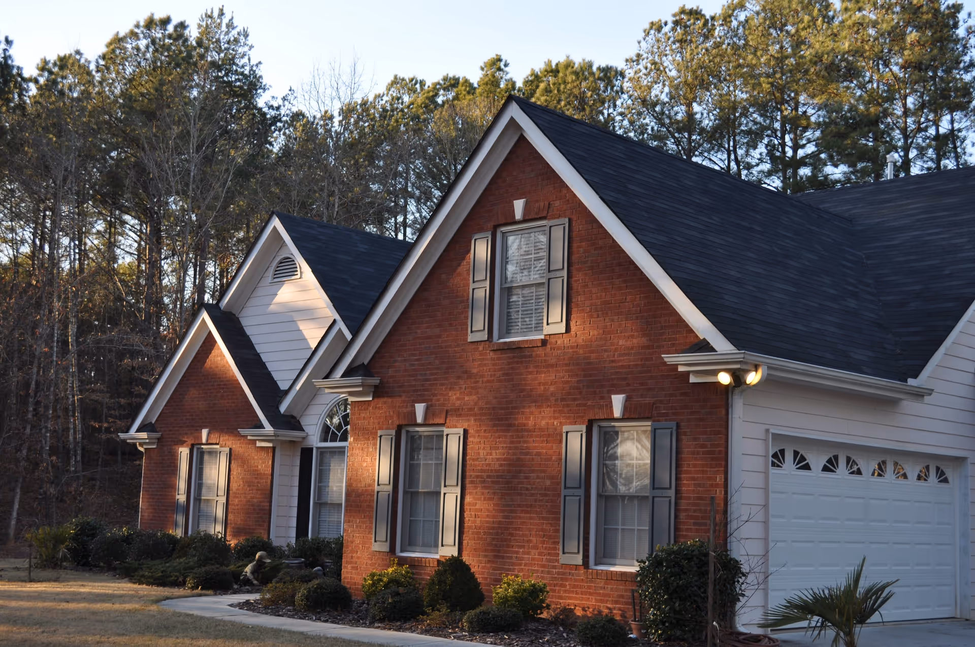 Exterior view of a single-story brick and siding house with a dark shingled roof, multiple windows with shutters, a white garage door, and surrounding landscaping including bushes and trees in the background.