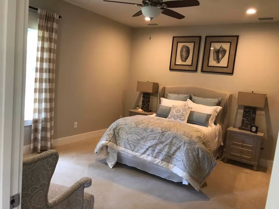 A neatly made bed with patterned bedding and multiple pillows in a bedroom. There are two matching nightstands on either side of the bed, each with a lamp. Two framed pictures hang above the bed. A ceiling fan with a light is mounted on the ceiling. A window with checkered curtains is visible on the left side, and a patterned armchair is partially visible in the foreground.