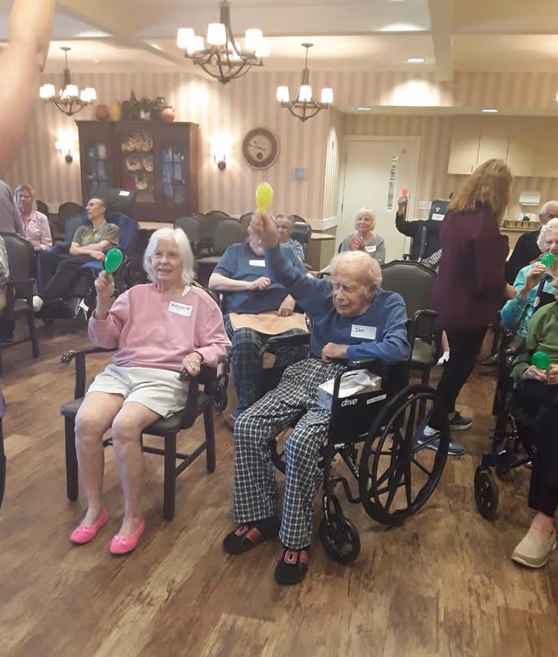A group of elderly people seated in a common room at a senior living facility, some holding colorful maracas and participating in an activity. The room has wooden floors, chandeliers, and a cabinet with decorative items in the background. One man is in a wheelchair, and a staff member is standing nearby.