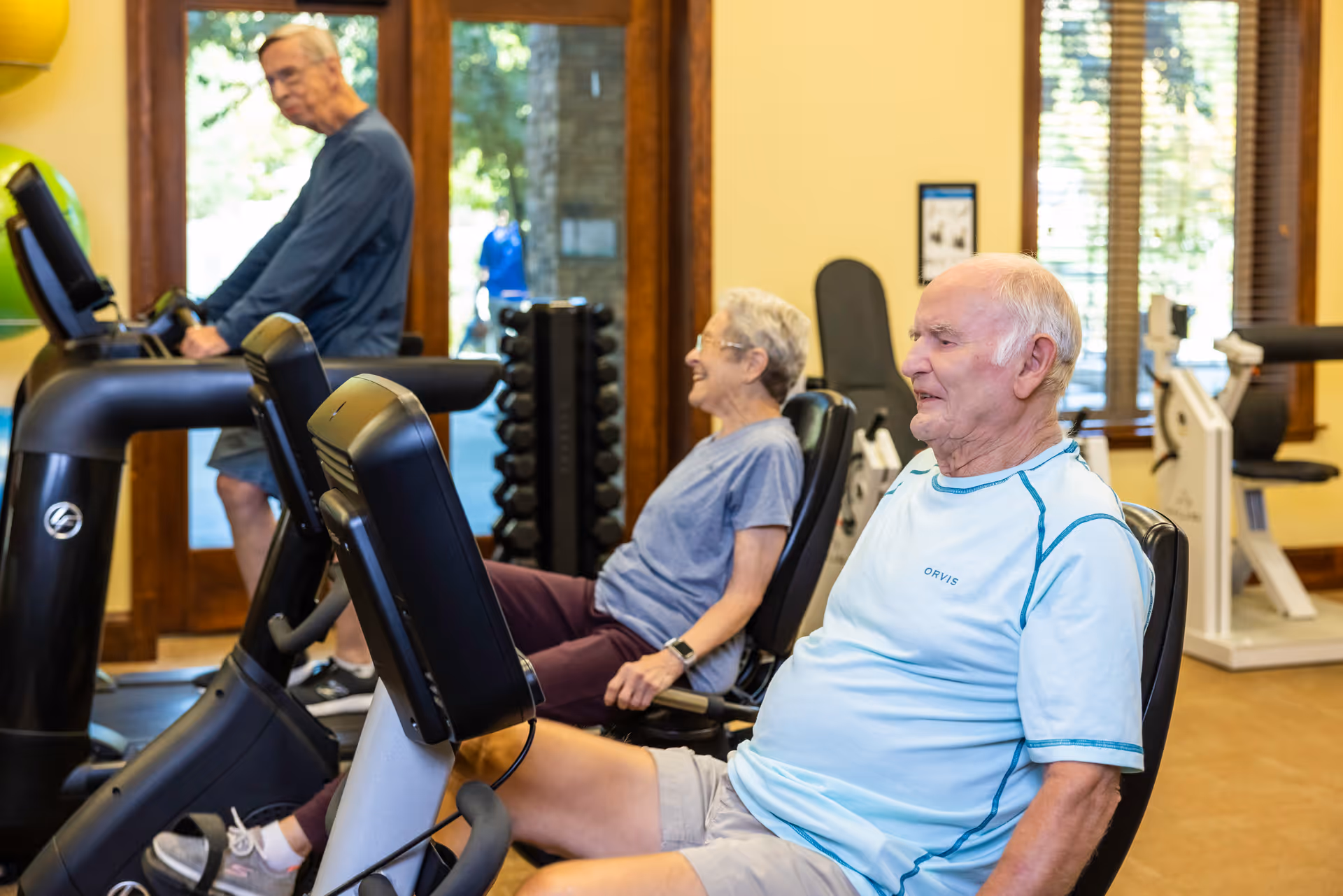 Three elderly individuals exercising on stationary bikes in a fitness room with large windows and wooden trim. The room has exercise equipment and natural light coming through the windows.