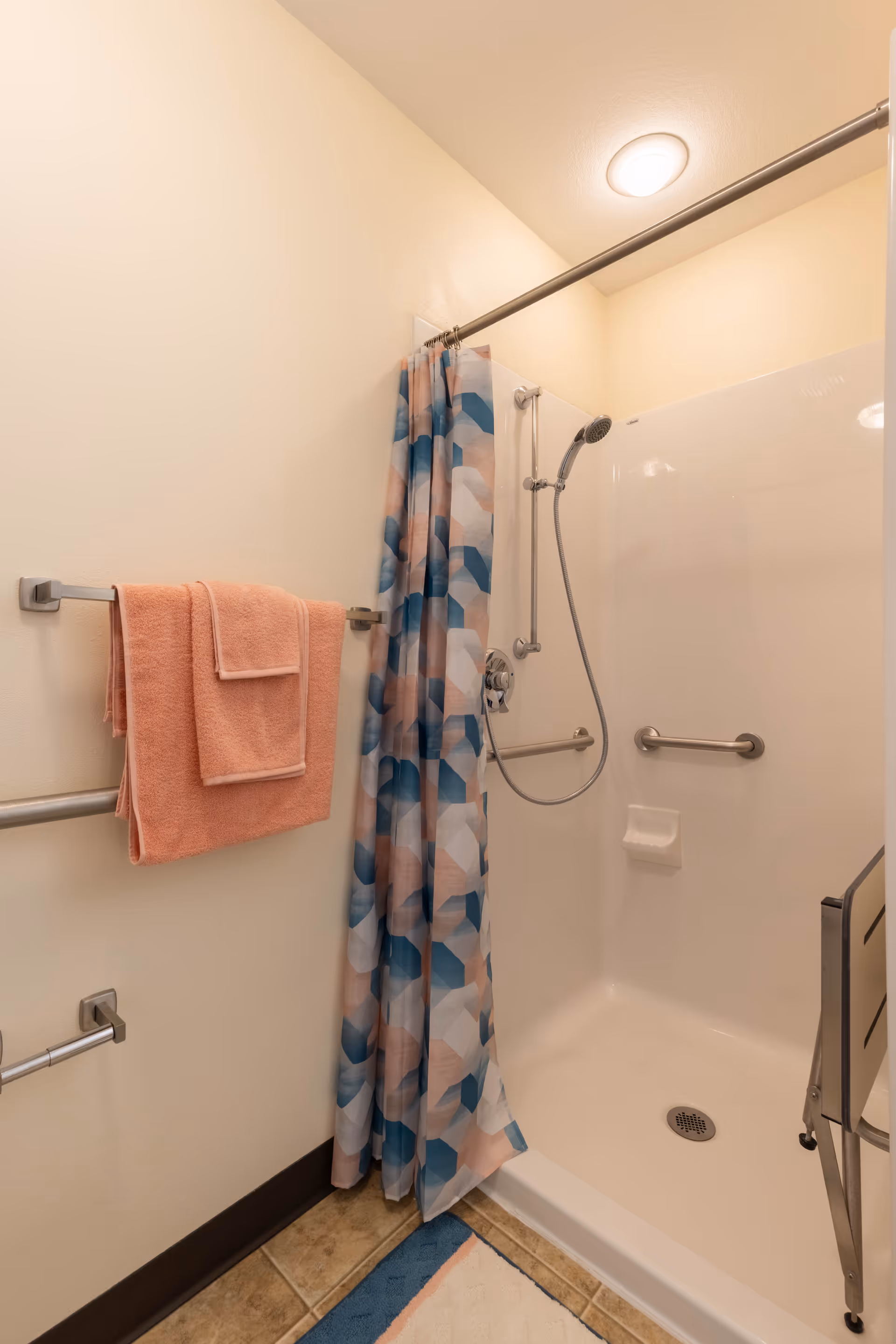 A shower area with a handheld showerhead, grab bars, and a colorful geometric-patterned shower curtain. Two peach-colored towels hang on a towel rack on the wall. The floor has a tiled surface with a blue and white bath mat.