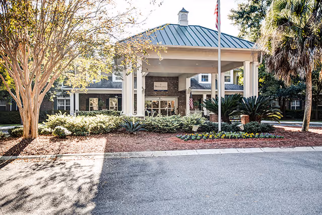 Front entrance of a residential building featuring a covered porte-cochere, flagpole, and landscaped flower beds.