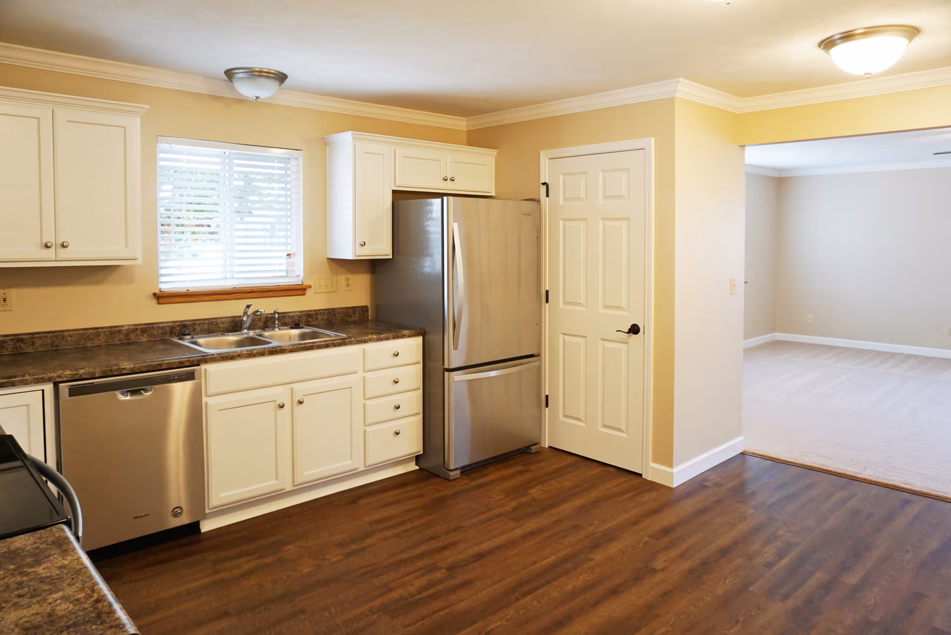A clean kitchen with white cabinets, a double sink under a window with blinds, a stainless steel dishwasher, and a stainless steel refrigerator next to a white door. The kitchen has brown countertops and wood flooring, with an adjacent room visible through an open doorway.