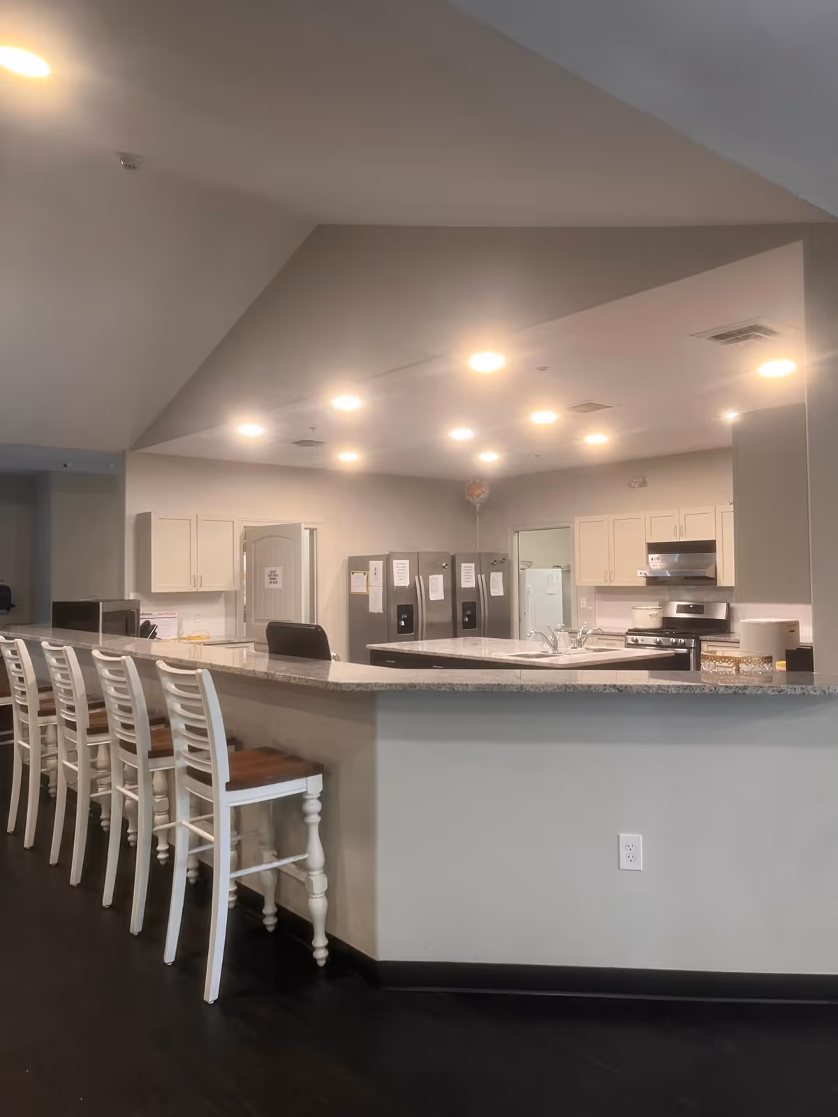 Communal kitchen area with a long marble-topped bar lined with white stools, multiple refrigerators, cabinets, and a stainless-steel stove.
