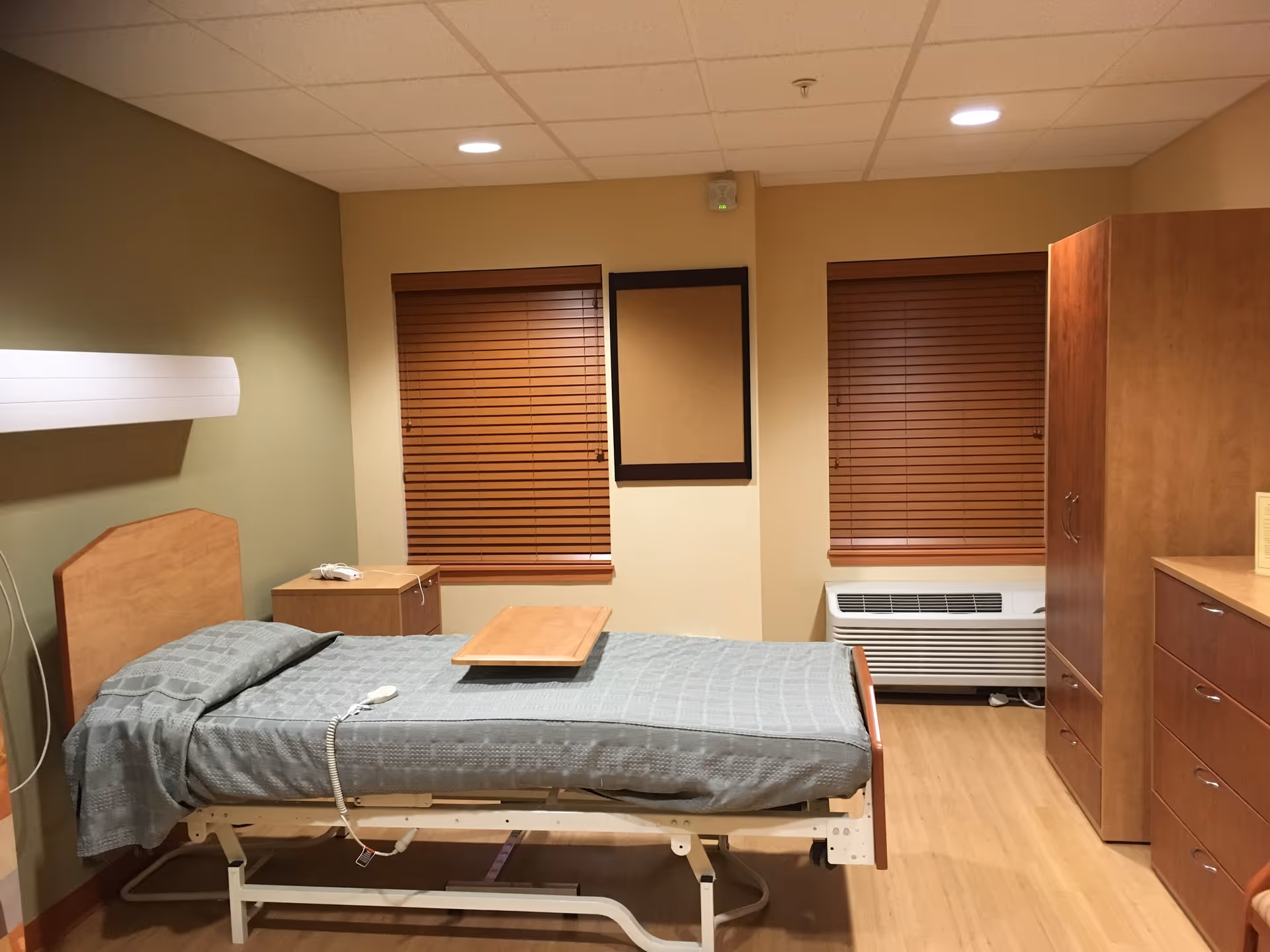 A simple furnished resident bedroom with a hospital-style bed, wooden furniture, and closed blinds.