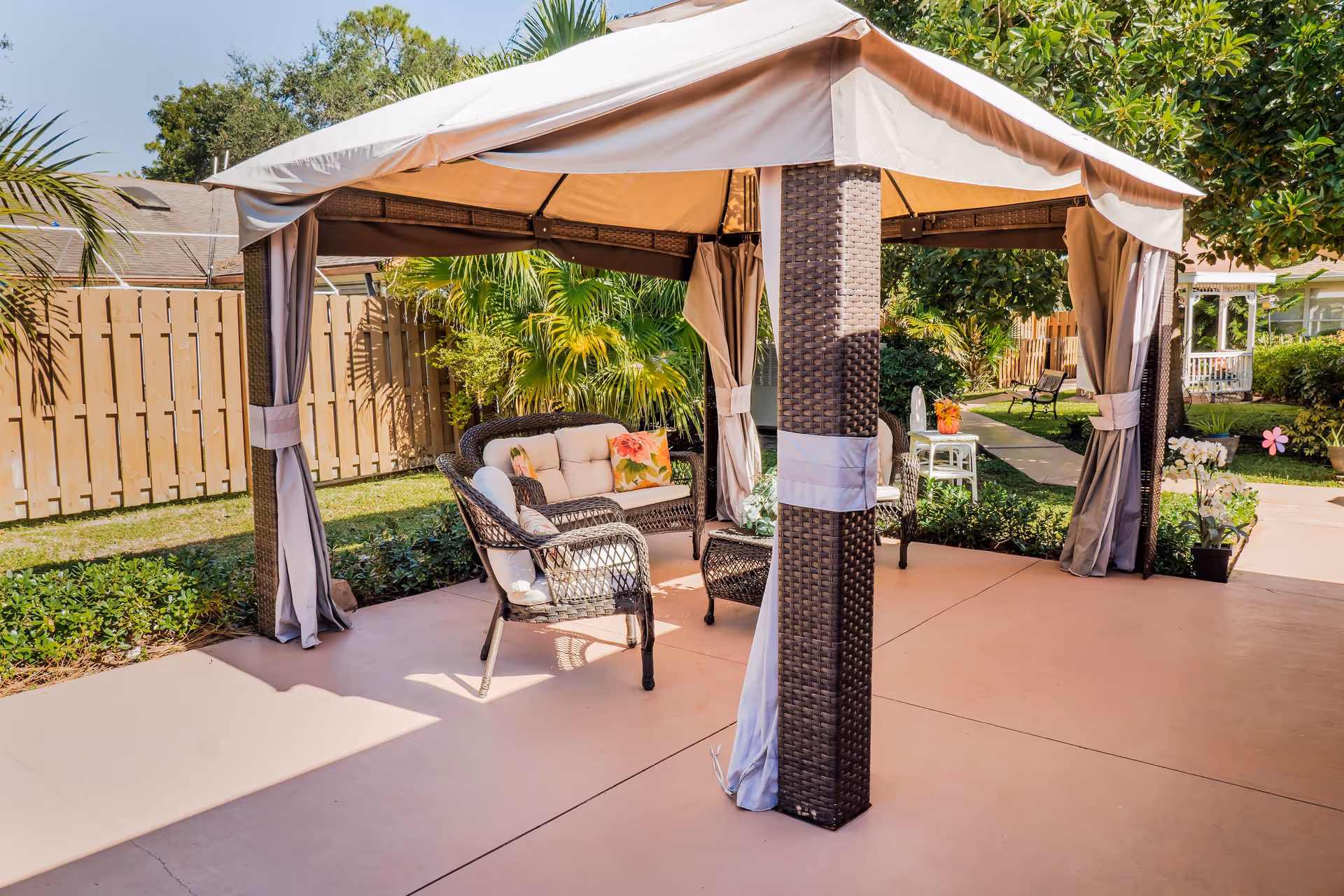 Outdoor patio area with a gazebo featuring a beige canopy and wicker pillars. Inside the gazebo, there are cushioned wicker chairs and a loveseat with floral pillows, surrounded by greenery and a wooden fence in the background. A paved walkway and garden benches are visible nearby.