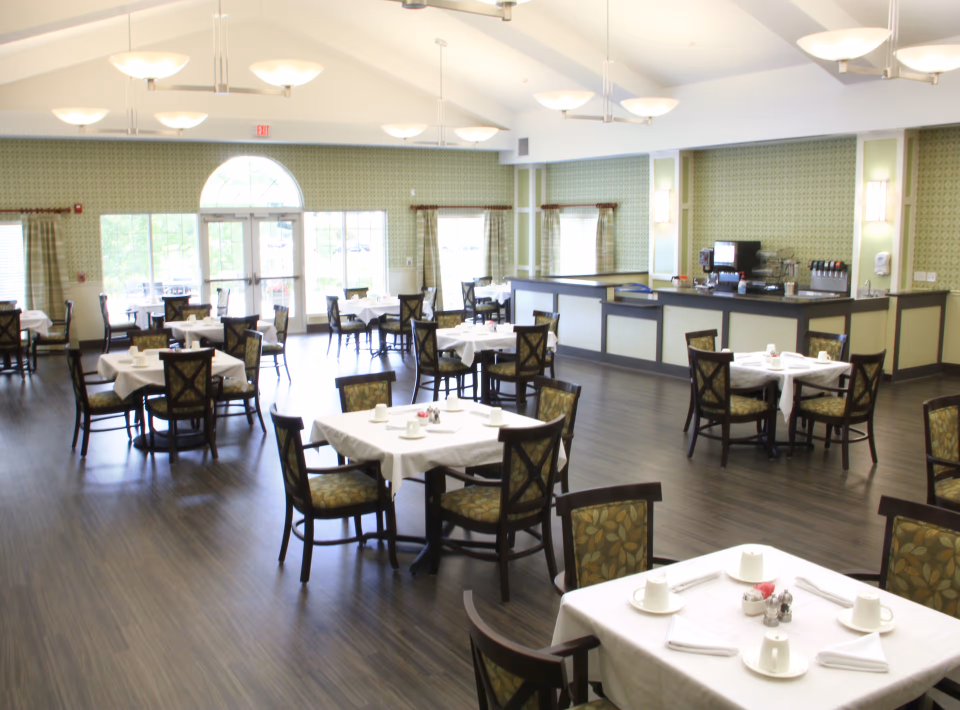 Sunlit dining room with multiple tables set for meals and a serving counter along the back wall.
