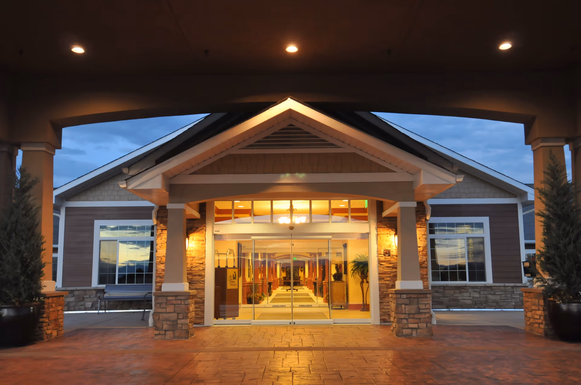 Entrance of a senior living facility with a covered driveway, stone pillars, large glass doors, and warm interior lighting visible inside during dusk.