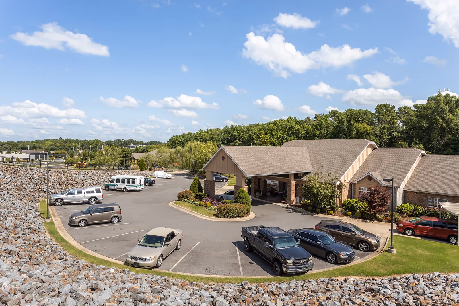 Exterior view of a single-story assisted living facility with a covered entrance, parking lot with cars and a small shuttle, and landscaped grounds under a blue sky.