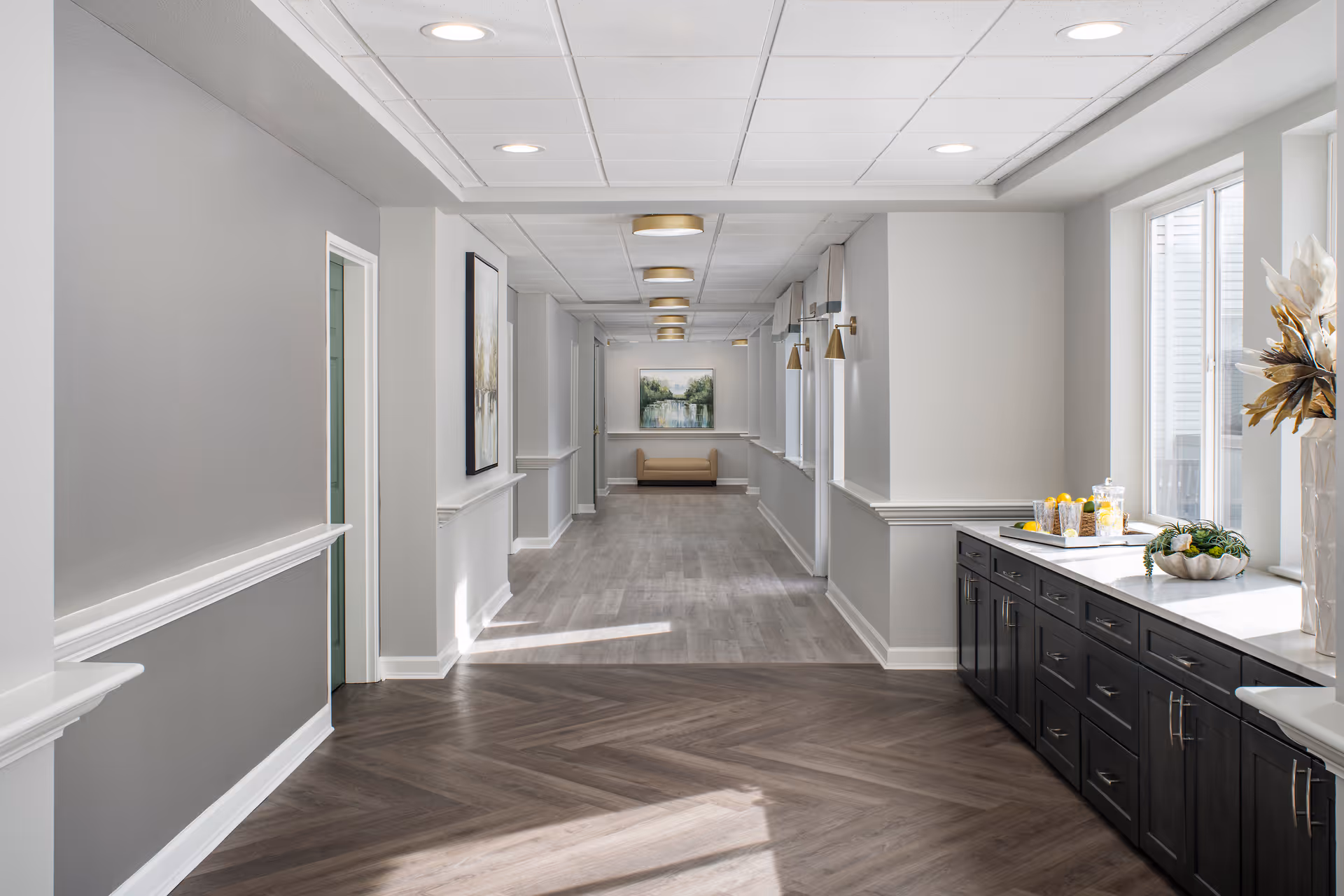 A bright, modern hallway in an assisted living facility with light gray walls, wood-patterned flooring, and recessed ceiling lights. On the right side, there is a long countertop with dark cabinets underneath, decorated with a vase of dried flowers, a bowl of greenery, and a tray with glasses and snacks. At the end of the hallway, a beige bench is positioned below a framed landscape painting.