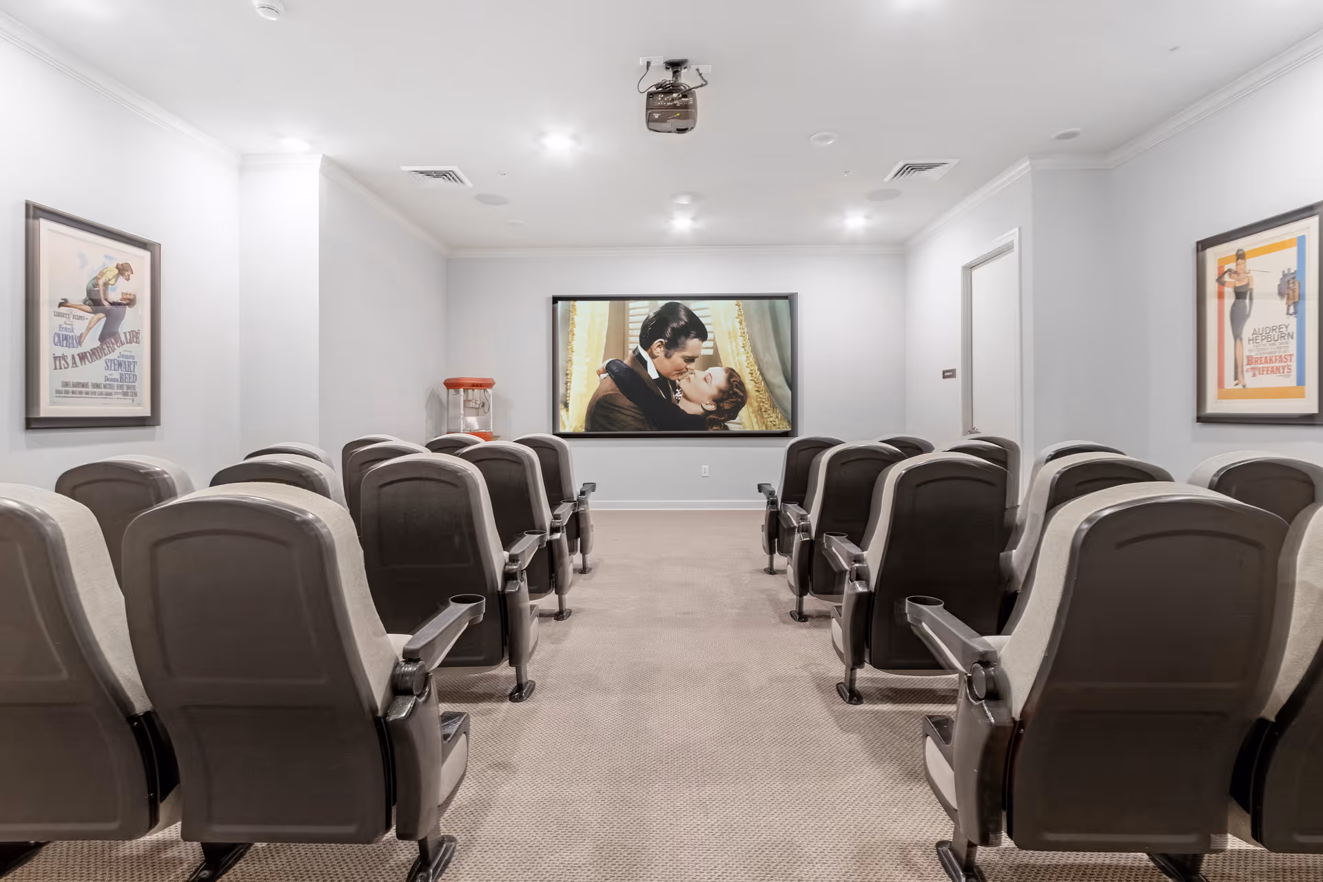 Small movie theater room with two rows of theater-style seats facing a large screen showing a classic movie scene. The walls are white with framed vintage movie posters on each side and a popcorn machine in the back corner.