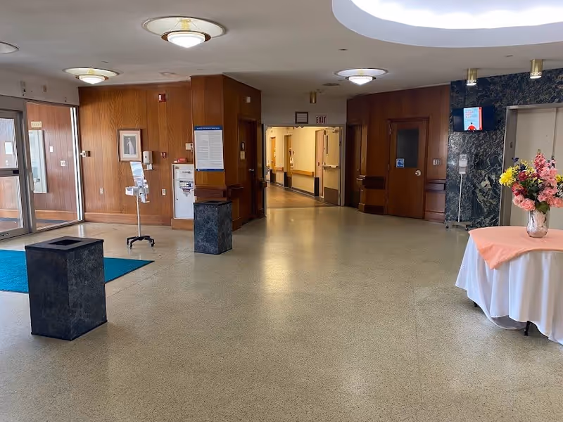Interior view of a senior living facility lobby with a polished floor, wooden panel walls, ceiling lights, a table with a pink tablecloth and a vase of flowers, and a hallway leading to other rooms. There are two dark stone trash bins and glass entrance doors on the left side.
