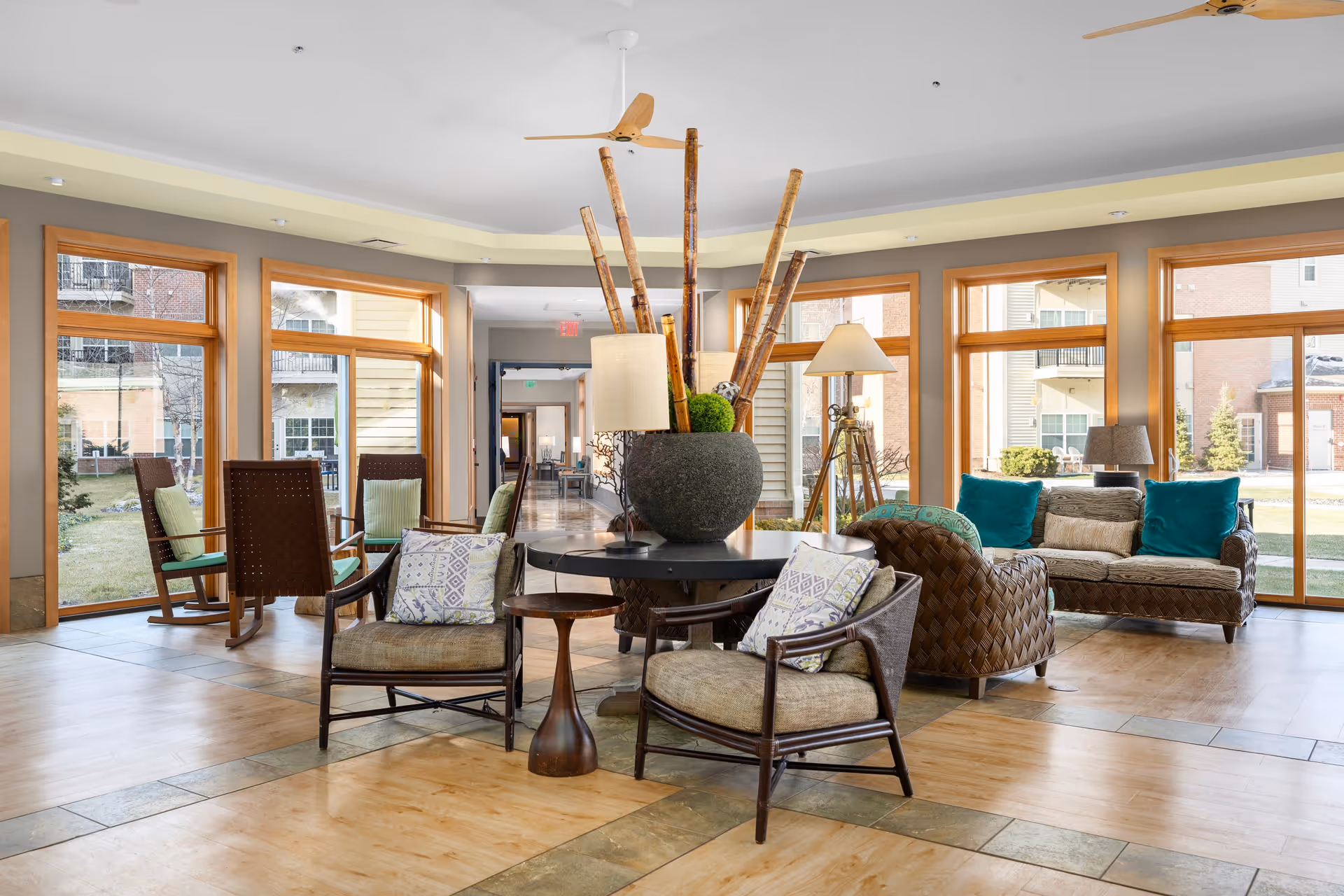 A bright and spacious living room area with large windows letting in natural light. The room features several chairs with cushions, a sofa with teal pillows, a round table with a large decorative vase holding tall bamboo sticks, and a floor lamp. The floor is wooden with tile accents, and outside the windows, parts of a garden and neighboring buildings are visible.