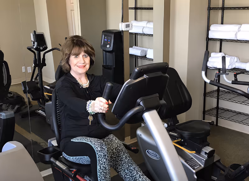 An elderly woman with short brown hair is sitting on a recumbent exercise bike in a fitness room. She is smiling and wearing a black top and patterned pants. Behind her, there are shelves with neatly folded white towels and other exercise equipment visible.