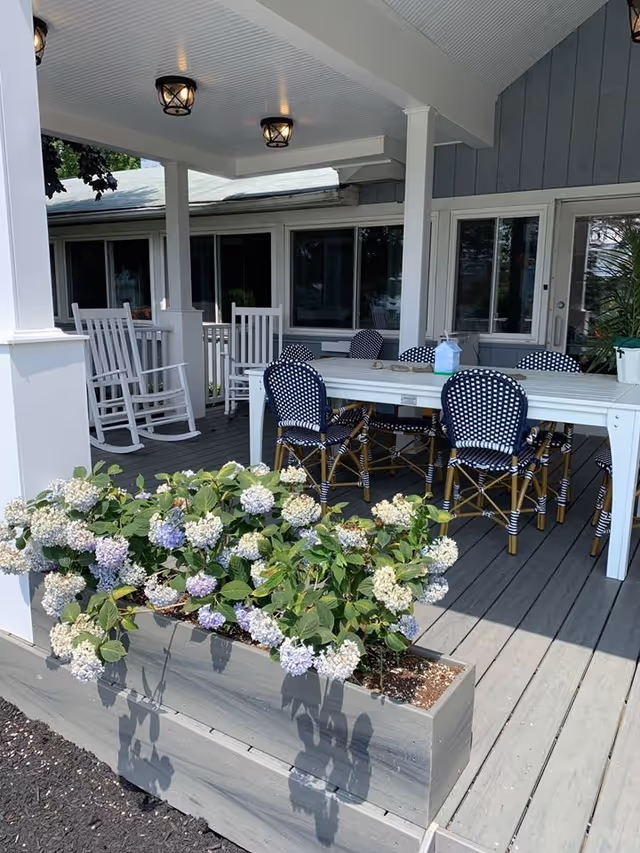 Covered outdoor patio area with a white table surrounded by six black and white woven chairs. There are two white rocking chairs in the background near the windows of the building. A planter box filled with blooming hydrangea flowers is in the foreground. The patio has a wooden floor and ceiling with three ceiling lights.