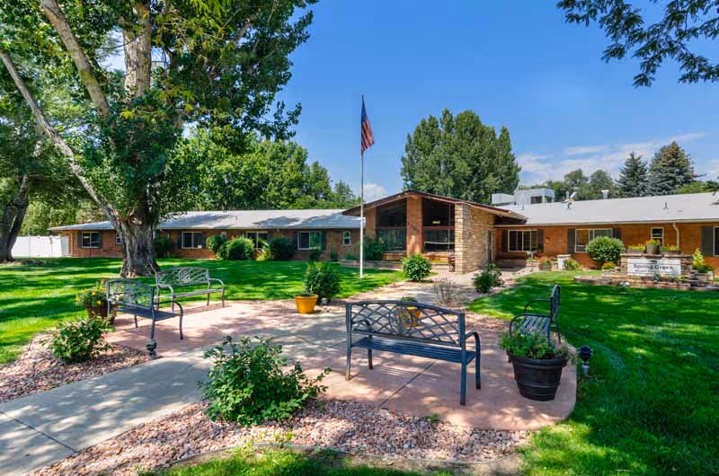 Landscaped courtyard with benches, a flagpole, and potted plants in front of a single-story brick nursing facility.