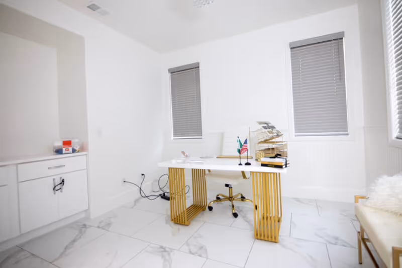 A bright, modern office space with white walls and marble tile flooring. The room features a white desk with gold legs, a white office chair with gold accents, two windows with closed blinds, a small cabinet with drawers on the left, and a beige chair with a white fluffy cushion on the right. On the desk are small flags, a book, and an organizer.