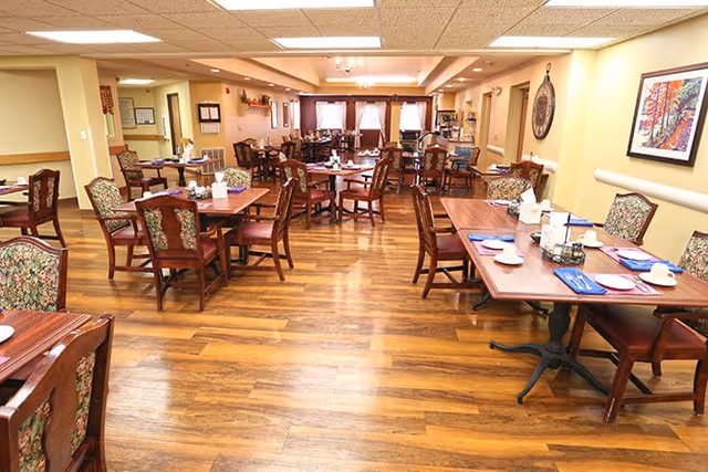 A spacious dining room with multiple wooden tables and chairs arranged neatly. The tables are set with plates, cups, napkins, and condiments. The room has warm lighting, wooden flooring, and light-colored walls adorned with framed artwork. Windows with curtains are visible at the far end of the room.