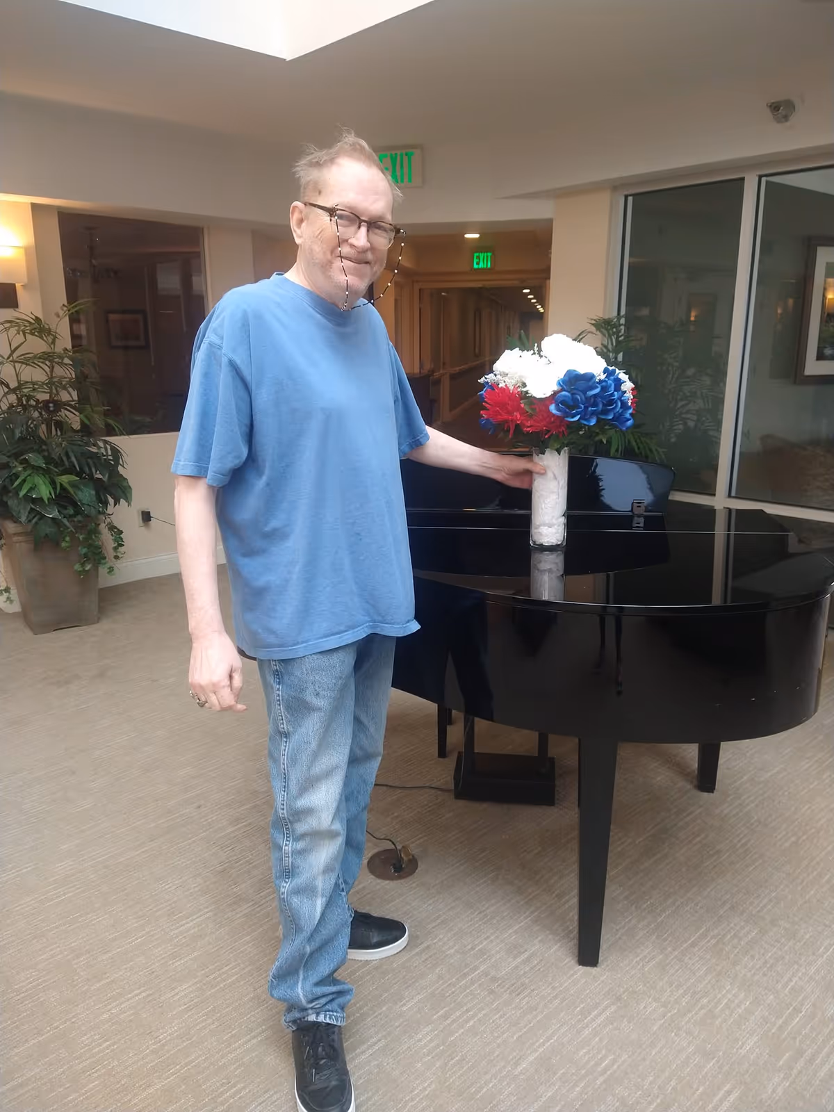 An elderly man wearing a blue t-shirt and jeans stands next to a black grand piano in a well-lit indoor area. He is holding a vase with red, white, and blue flowers placed on the piano. There are plants and framed pictures in the background, along with visible exit signs.