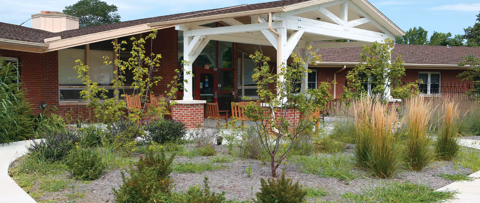 Single-story brick nursing facility with a white covered entrance, rocking chairs on the porch, and landscaped garden beds and walkways in front.