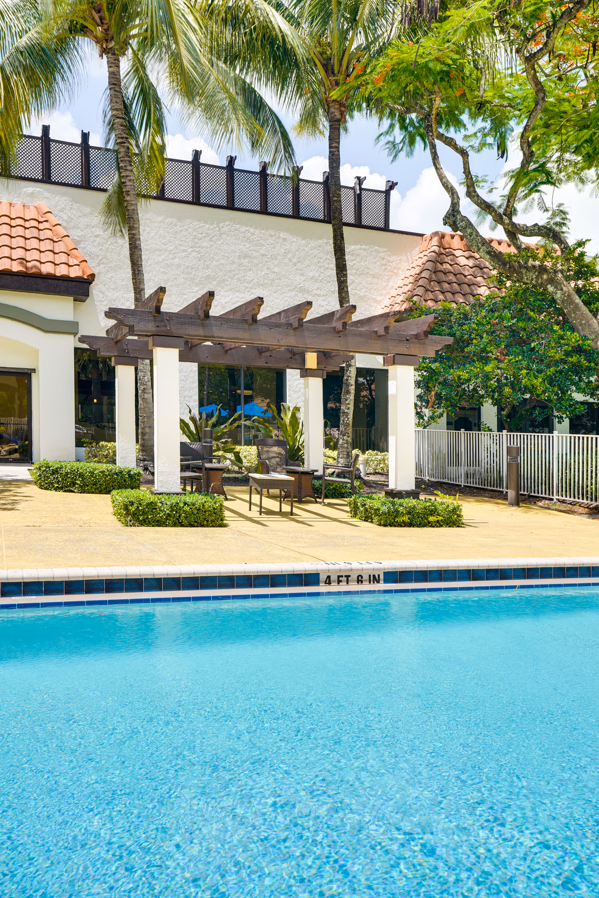 Outdoor swimming pool with clear blue water in front of a building with a tiled roof. There is a shaded seating area with a wooden pergola, chairs, and a table surrounded by greenery and palm trees.